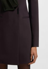 Cappotto su misura viola con una texture liscia, caratterizzato da un accento nero al colletto e nelle tasche laterali. Maniche lunghe e design elegante.