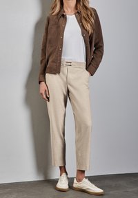 Hnědá semišová bunda přes bílé tričko, kombinovaná s béžovými kalhotami na míru a bílými teniskami s opálenými podrážkami, na šedém pozadí.