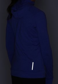 Giacca con cappuccio in tessuto blu scuro con un aspetto testurizzato. Presenta un dettaglio riflettente vicino all'orlo e linee di cucitura ergonomiche.