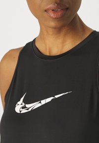 Czarny sportowy top bez rękawów z białym logo Nike z abstrakcyjnymi detalami; gładka tkanina i okrągły dekolt.