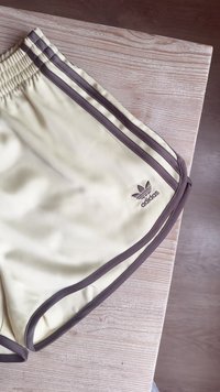 Calções atléticos amarelos com riscas laterais castanhas, cintura elástica e acabamento castanho. Apresenta o logo da Adidas bordado na parte inferior esquerda.