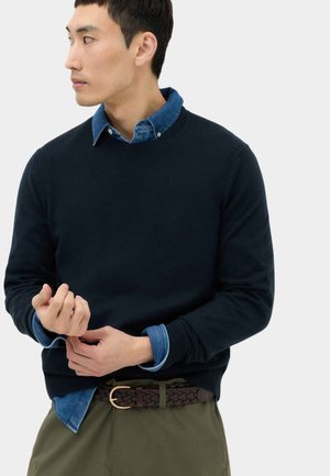 Mand iført en mørk sweater over en blå skjorte med knapper, justerer sin ærme, iført olivengrønne bukser og et fletbælte af læder.