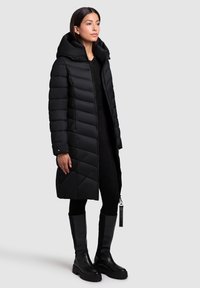 Black padded coat with a high collar and hood, featuring quilted texture and a front zipper. Paired with knee-high black boots.
