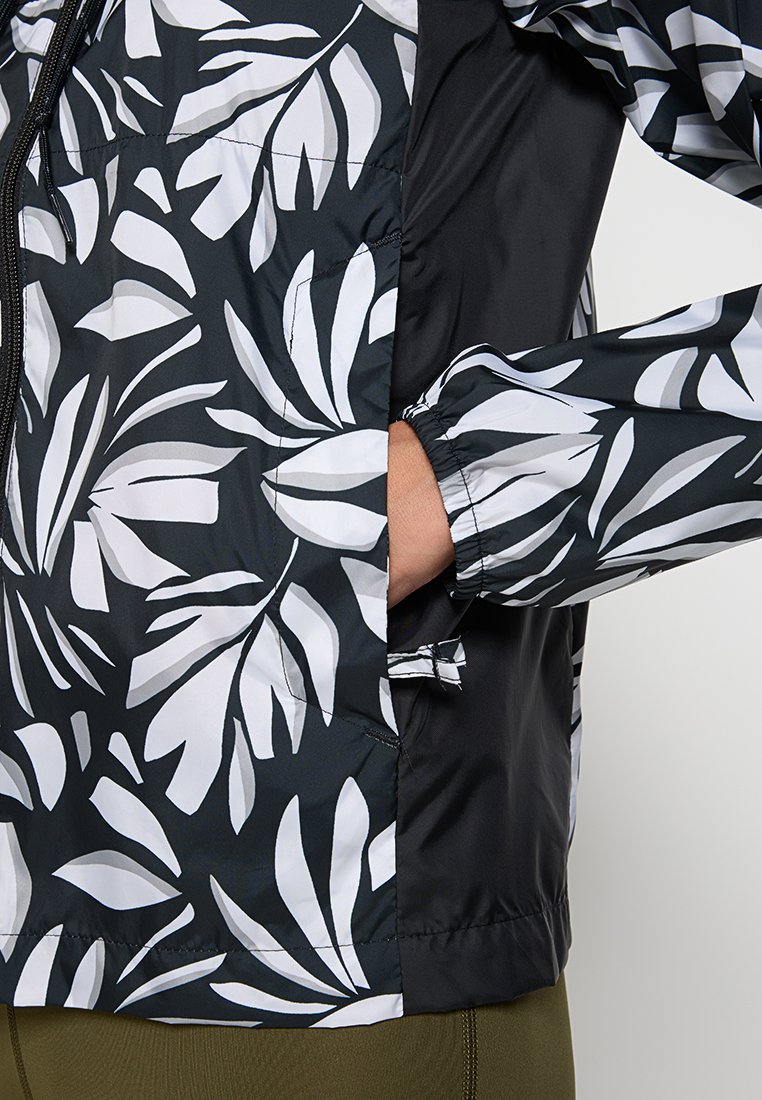 Chaqueta con estampado de hojas en blanco y negro, con cremallera frontal y puños elásticos, mostrando una mano dentro de un bolsillo lateral.