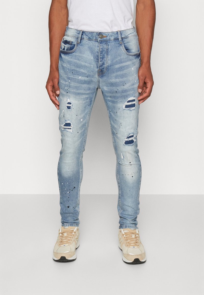 Faded blue denim jeans with ripped details, paint splatter accents, and a slim fit design. Front pockets included.