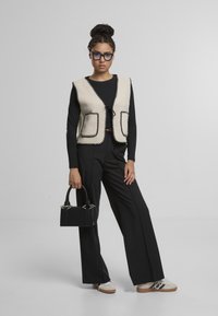 Vesta z vlněné směsi v krémové barvě s černými detaily, s předními kapsami a zavazováním na mašli. Černý top s dlouhým rukávem, laděné černé kalhoty a strukturovaná černá kabelka.
