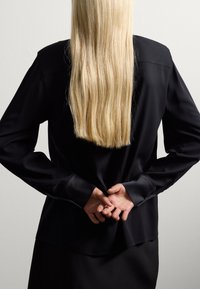 Black, long-sleeved shirt with a smooth texture and a loose fit, worn with hands clasped behind the back. Long, straight blonde hair is visible.