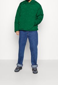 Giacca trapuntata verde con colletto, abbinata a jeans blu con risvolti e sneakers grigie. La giacca presenta un motivo a rombi.