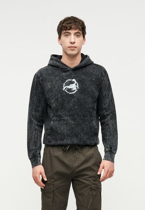 Jeune homme portant un sweat à capuche noir délavé à l'acide avec un logo de homard blanc et du texte, associé à un pantalon cargo olive, se tenant devant un fond uni.