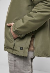 Olivi vihreä takki, jossa on sileä pinta, vetoketjullinen sivutasku ja kangaslogotagi helmassaan. Vaatteena ruskea paita ja harmaat housut.