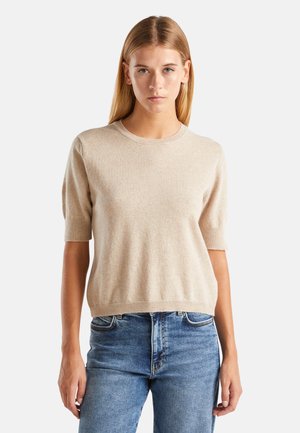Young woman with straight blonde hair wearing a beige short-sleeve sweater and blue jeans standing against a plain background.