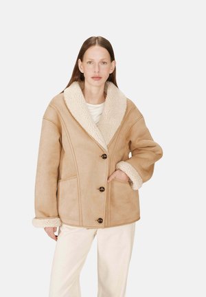 Manteau en shearling beige avec un grand col, deux poches frontales et trois boutons foncés. Texture douce et design surdimensionné.