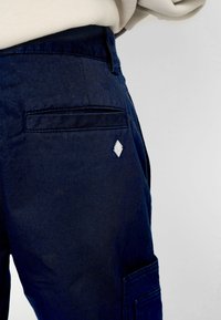 Los pantalones cargo de mezclilla azul marino cuentan con un bolsillo trasero, un acento metálico en forma de diamante y un corte recto con una textura suave.