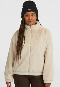 Beige fleece jas met hoge kraag, ritssluiting aan de voorkant en elastische manchetten. Draagt over een donker shirt, gecombineerd met een zwarte gebreide beanie.