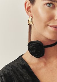 Zlato-tonirane abstraktne uhane dopolnjuje črn žametni ovratnik z dizajnom vrtnice. Oblačilo je temno in iz teksturirane tkanine.