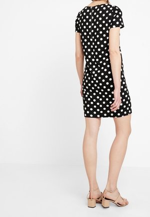 Femme portant une robe noire à manches courtes avec des pois blancs et des sandales à talons carrés beige, debout de dos sur un fond blanc.