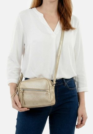 Vrouw draagt een witte blouse en donkere spijkerbroek, houdend een kleine metalen gouden crossbodytas met ritssluitingen.