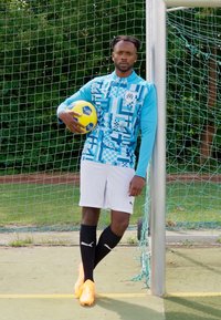 Maglia blu a maniche lunghe e a fantasia, pantaloni bianchi, calze nere e scarpe arancioni. Il giocatore tiene un pallone da calcio giallo vicino a un palo della porta.