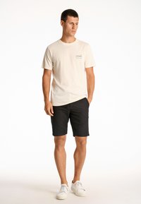 Vaaleanbeige puuvillapaita, jossa on pieni graafinen kuvio ja lyhyet hihat, yhdistetty mustiin shortseihin ja valkoisiin tennareihin. Yksinkertainen, rento malli.