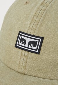 Khaki cap laget av teksturert stoff med en svart rektangulær lapp som har to hvite konturerte dyresilhuetter. Seks-panel design med søm.