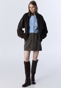 Vrouw die staat in een zwarte oversized jas, lichtblauwe gebuttonde cardigan, bedrukte minirok, lange donkerbruine laarzen en blauwe oorbellen.