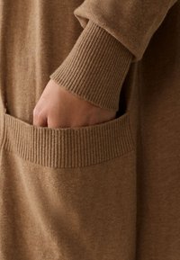Brown knitted garment with ribbed cuffs and a pocket. Material appears soft with a subtle texture, emphasizing comfort and warmth.