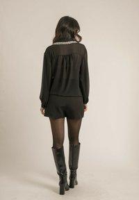 Haut noir transparent avec un col en perles, associé à des shorts noirs et des bottes noires jusqu'aux genoux avec un talon carré, créant une silhouette élégante.