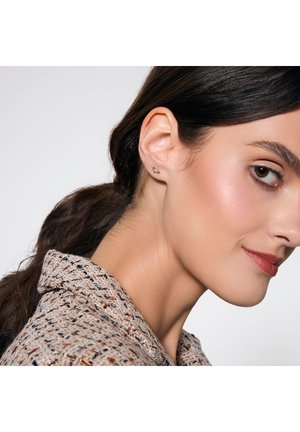 Złote kolczyki wkrętki z kwiatowym wzorem, z teksturowaną powierzchnią. Modelka ma na sobie wzorzystą bluzkę w neutralnych odcieniach z kołnierzykiem.