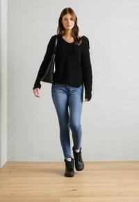 Svart strikkegenser med løs passform, blå skinny jeans og svarte støvler. Modellen bærer en svart håndveske. Treverk gulv og lys vegg som bakgrunn.
