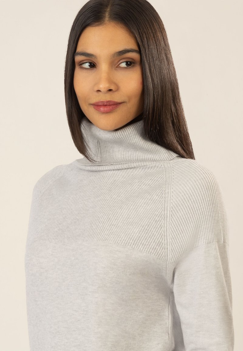 Hellgrauer Pullover mit geripptem Rollkragen, glatter Oberflächenstruktur und markanten Rippendetails an den Schultern.