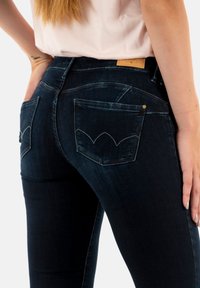 Jean skinny en denim noir avec des poches arrière arrondies, des coutures contrastées et un patch en cuir sur la taille.