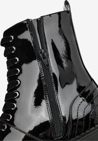 Botín de charol negro brillante con cremallera lateral y cordones negros atravesados por ojales de metal.