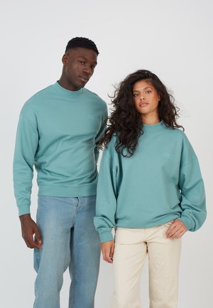 Tyrkise gensere med rund hals, ribbestrikkede mansjetter og matchende brodering, parret med lyse jeans og kremfargede bukser. Avslappet passform og myk tekstur.