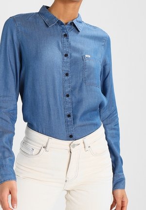 Persoon draagt een blauwe denim blouse met lange mouwen en knopen, ingestopt in een witte hoge taille jeans met voorzakken.