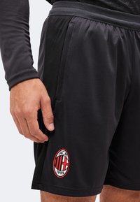Μαύρα αθλητικά σορτς με το λογότυπο της AC Milan στο αριστερό πόδι, φορεμένα από ένα άτομο με ορατό χέρι και μπράτσο που φοράει μαύρο μακρυμάνικο μπλουζάκι.