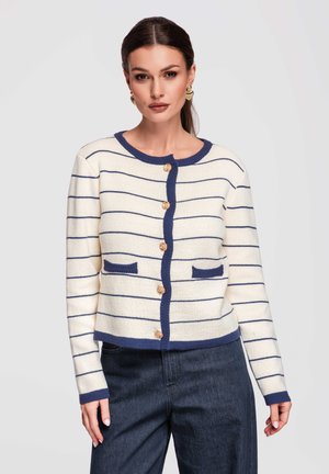 Kvinde iført cremefarvet strikket cardigan med mørkeblå horisontale striber, mørkeblå kant, guldfarvede knapper og mørkeblå jeans, stående foran en ensfarvet baggrund.