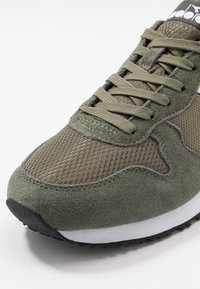 Zapatilla verde oliva con parte superior de malla y acentos de ante, puntera redondeada, diseño de cordones y suela negra de goma texturizada.
