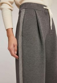 Grijze joggingbroek van zacht materiaal met een trekkoord in de taille, plooien aan de zijkant en witte strepen langs de zijkanten.