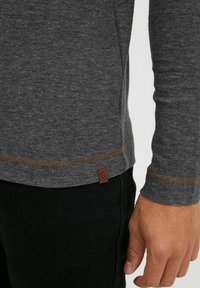 Graues Langarmshirt aus strukturiertem Stoff, mit einem braunen gesteppten Saum und einem kleinen Lederlogo-Anhänger an der unteren linken Seite.