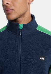 Μπλε φλις μπουφάν με ψηλό γιακά, πράσινες μαλακές λεπτομέρειες στους ώμους και φερμουάρ στο μπροστινό μέρος. Διαθέτει μικρό λογότυπο στο στήθος.