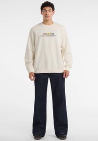 Młody mężczyzna w kremowym swetrze z napisem "Guess Jeans Company" i ciemnych szerokich spodniach, stojący na białym tle.