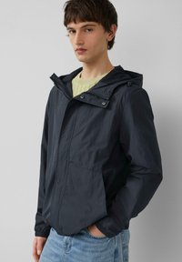 Lichte zwarte hoodie met een ritssluiting aan de voorkant, elastische manchetten en twee zijzakken. De stof heeft een gladde, licht glanzende textuur.