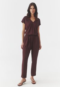 Bordeaux jumpsuit van zachte stof, met een V-hals, korte mouwen en een elastische tailleband met strik, gecombineerd met sandalen met dunne bandjes.