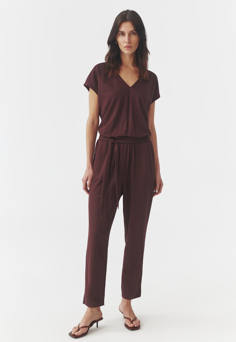 Bordeaux jumpsuit van zachte stof, met een V-hals, korte mouwen en een elastische tailleband met strik, gecombineerd met sandalen met dunne bandjes.