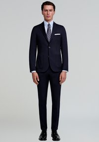 Traje azul marino oscuro con chaqueta de un solo botón, solapas con muesca y dos bolsillos frontales. Combinado con una camisa clara y una corbata de estampado. Zapatos negros.