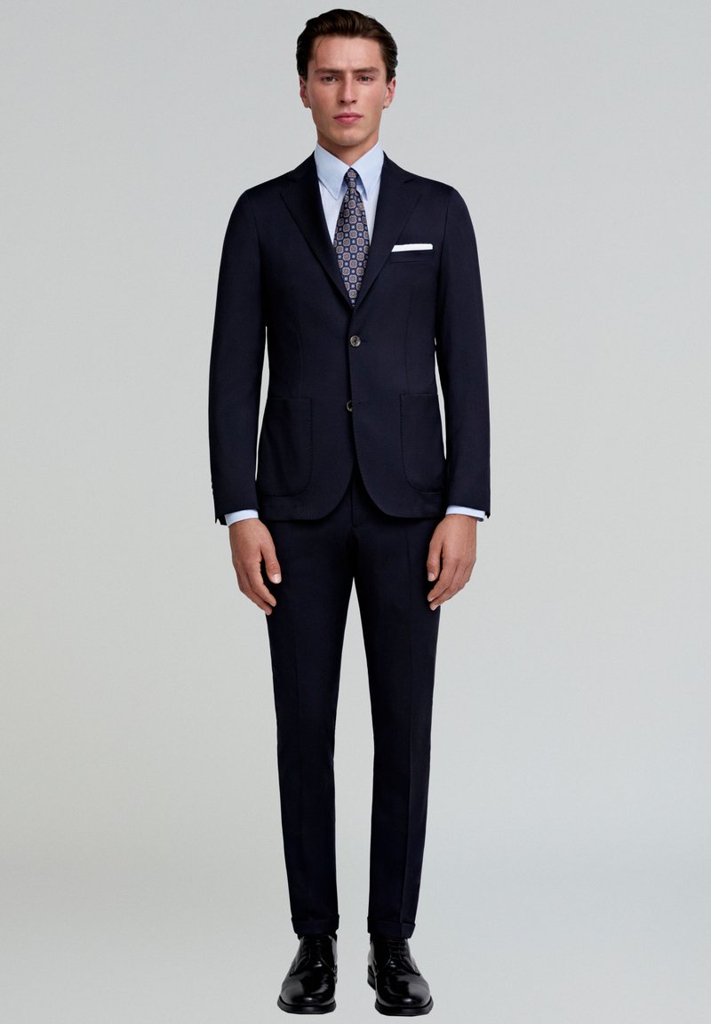 Traje azul marino oscuro con chaqueta de un solo botón, solapas con muesca y dos bolsillos frontales. Combinado con una camisa clara y una corbata de estampado. Zapatos negros.