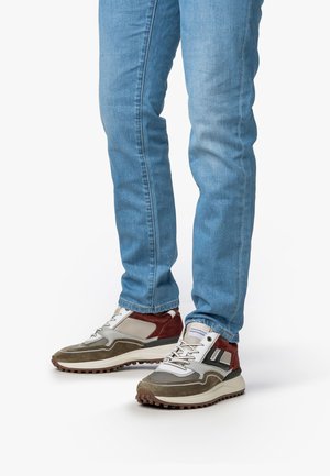Des jambes en jeans bleu clair portant des baskets marron, beige et blanches avec des semelles texturées sur un fond blanc.