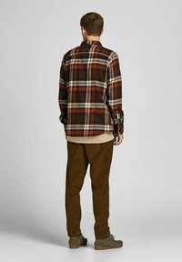 Camicia di flanella a quadri nei colori marrone, arancione e crema, con polsini abbottonati e colletto, indossata sopra un top beige a strati e pantaloni verde oliva.