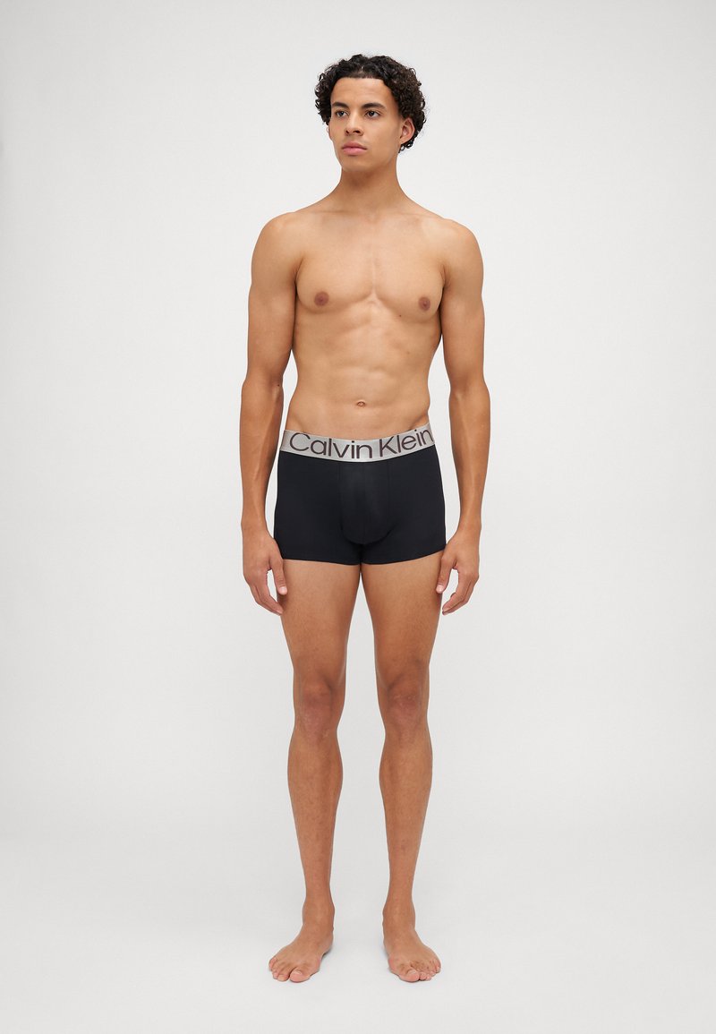 Črne bombažne bokserice z sivo pasom s Calvin Klein logotipom, ki imajo prilagojen kroj in gladko teksturo. Model stoji bos.