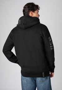 Schwarzer Hoodie aus weichem Stoff, mit großer Kapuze, gerippten Bündchen und einem Logo, das auf dem Rücken und dem Ärmel gedruckt ist. Kombiniert mit Jeans.